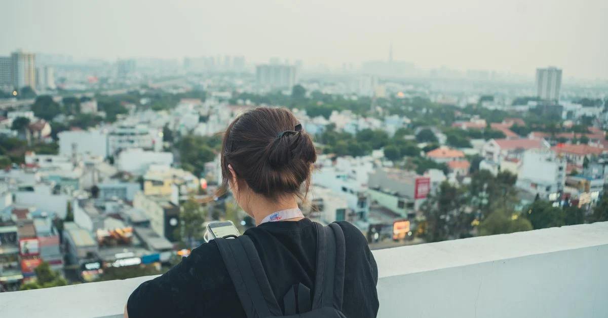Une femme avec un sac à dos se tient sur un toit, utilisant son téléphone pour capturer la vue urbaine.