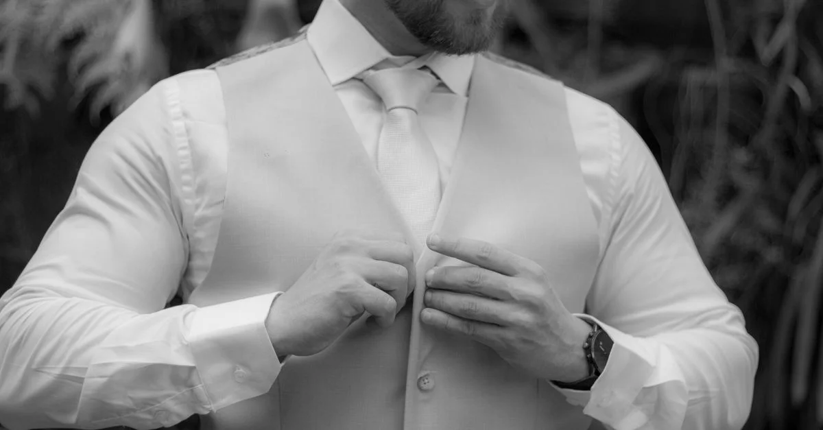 Un homme sophistiqué qui redresse son gilet, dégageant style et confiance dans un décor monochrome.