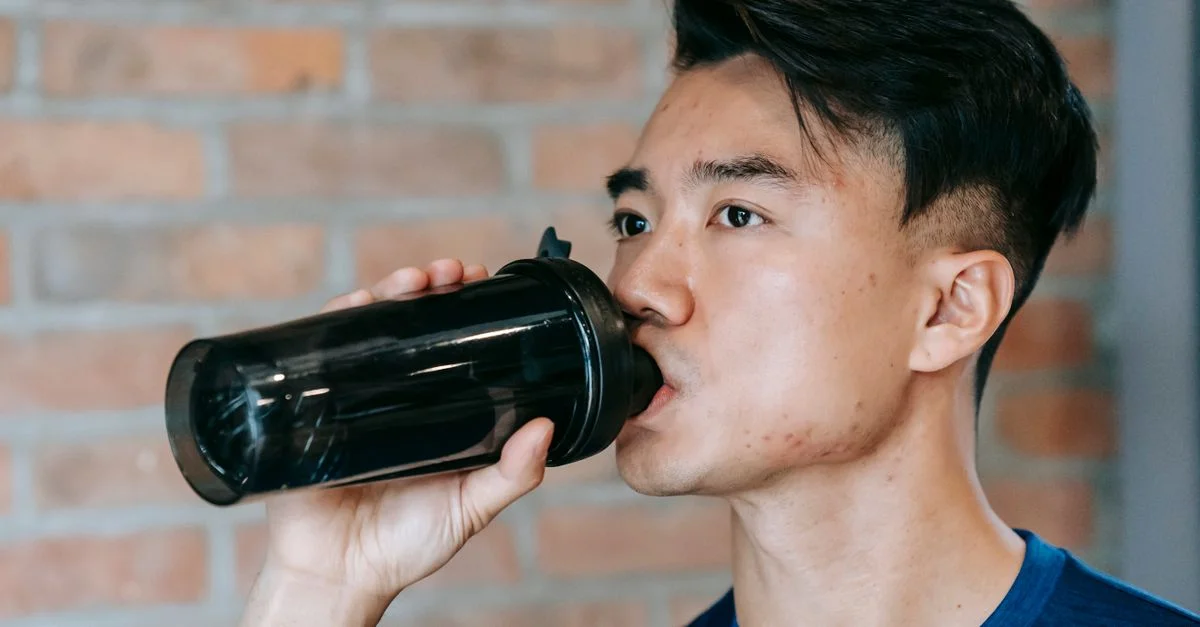Jeune homme buvant de l'eau d'une bouteille shaker dans une salle de sport, restant concentré et hydraté.